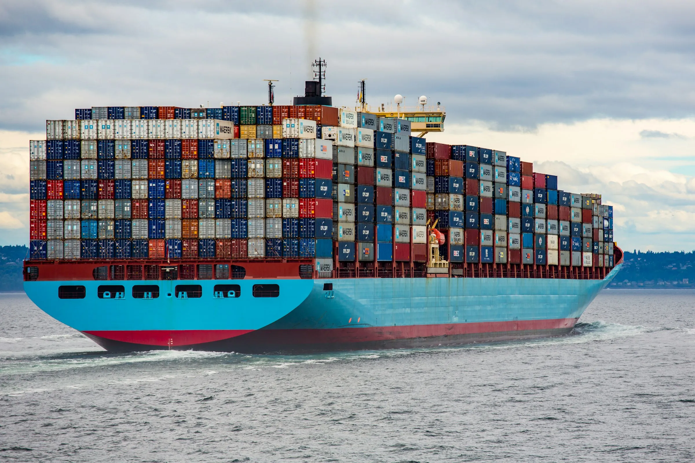 A massive container ship with stacked cargo containers sailing across the ocean under a clear blue sky.
